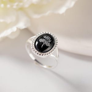 K&ouml;nnte beinhalten: Ein silberner Ring mit einem ovalen, schwarzen Stein mit floralem Design. Der Ring hat einen dekorativen Rand um den Stein und ein verstellbares Band. Der Hintergrund ist verschwommen, mit einer wei&szlig;en Blume sichtbar.