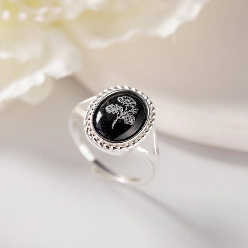 K&ouml;nnte beinhalten: Ein silberner Ring mit einem ovalen, schwarzen Stein mit floralem Design. Der Ring hat einen dekorativen Rand um den Stein und ein verstellbares Band. Der Hintergrund ist verschwommen, mit einer wei&szlig;en Blume sichtbar.