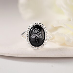 K&ouml;nnte beinhalten: Silberring mit einem ovalen, schwarzen Stein mit floralem Design. Der Stein ist in einem geflochtenen Silberrahmen gefasst. Das Band des Rings teilt sich in zwei zarte Silberstr&auml;nge auf jeder Seite des Steins.