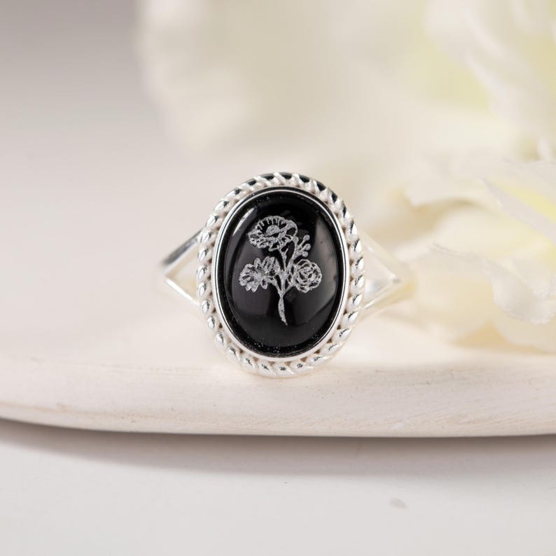 K&ouml;nnte beinhalten: Silberring mit einem ovalen, schwarzen Stein mit floralem Design. Der Stein ist in einem geflochtenen Silberrahmen gefasst. Das Band des Rings teilt sich in zwei zarte Silberstr&auml;nge auf jeder Seite des Steins.
