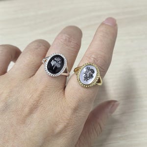 K&ouml;nnte beinhalten: Zwei Ringe mit ovalen Gesichtern, einer silbernen und einer goldenen. Der silberne Ring hat ein schwarzes Gesicht mit einem wei&szlig;en Blumendesign, w&auml;hrend der goldene Ring ein wei&szlig;es Gesicht mit einem Blumenstrau&szlig; aufweist. Beide Ringe sind an Fingern.