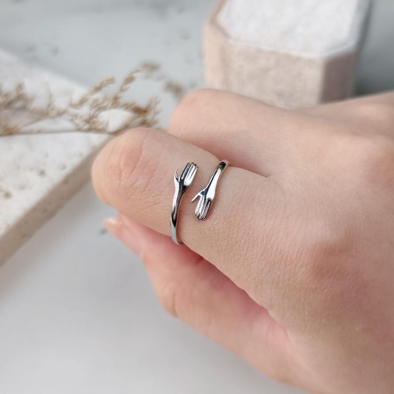 Może przedstawiać: Srebrny pierścionek z dwoma splecionymi dłońmi, tworzącymi okrąg. Pierścionek noszony jest na palcu.