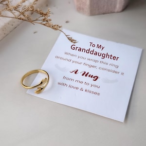 Puede incluir: Un anillo de metal dorado con forma de dos manos que se abrazan, colocado sobre una tarjeta blanca con el texto impreso: To My Granddaughter, When you wrap this ring around your finger, consider it A Hug from me to you with love & kisses.