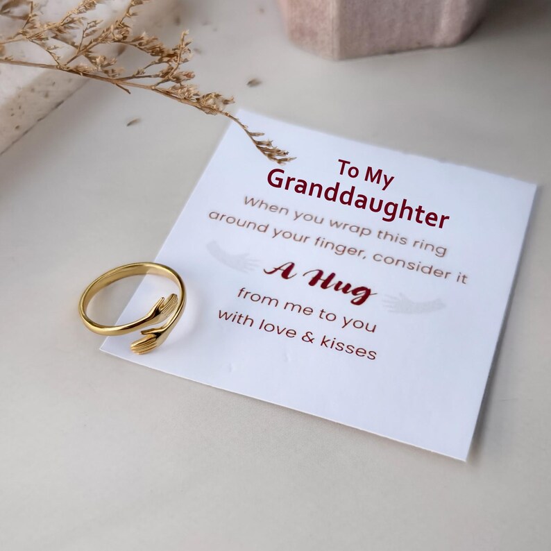 Puede incluir: Un anillo de metal dorado con forma de dos manos que se abrazan, colocado sobre una tarjeta blanca con el texto impreso: To My Granddaughter, When you wrap this ring around your finger, consider it A Hug from me to you with love & kisses.