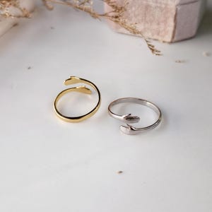 Puede incluir: Dos anillos abiertos, uno en tono dorado y otro en tono plateado, cada uno con un dise&ntilde;o de dos manos que se encuentran en el centro para formar un abrazo. Las bandas son delgadas y circulares, con los detalles de las manos visibles.
