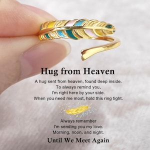 以下が含まれることがあります： 金色の羽の指輪、カラフルなエナメル装飾。リングは黄色、ピンク、青、ターコイズのアクセント。画像には「Hug from Heaven」というテキストと、愛と記憶に関するメッセージが含まれています。