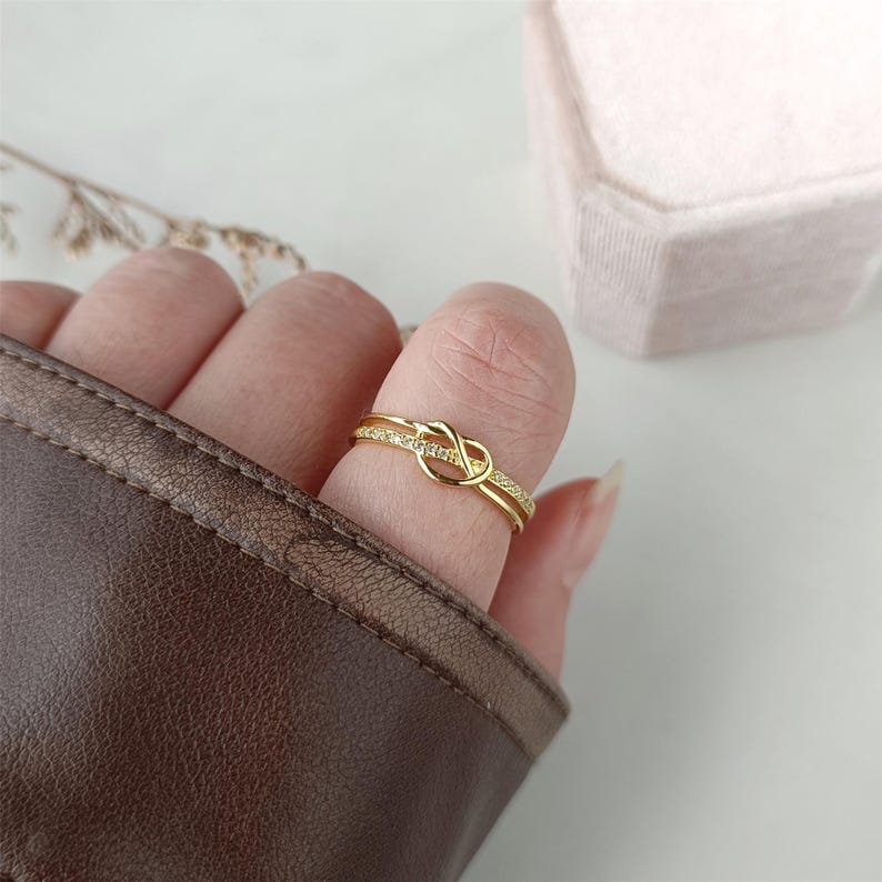 May include: A gold ring with a knot design and small clear stones. The ring is on a finger.