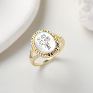 K&ouml;nnte beinhalten: Goldfarbener Ring mit einem ovalen Mittelteil aus Perlmutt. Das Zentrum zeigt ein schwarz-wei&szlig;es Blumendesign. Der Ring hat einen geflochtenen Rand und ein geteiltes Schaftdesign.