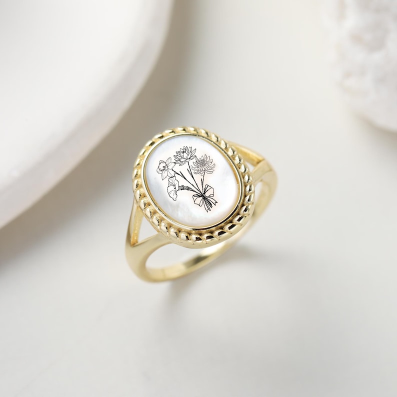 K&ouml;nnte beinhalten: Goldfarbener Ring mit einem ovalen Mittelteil aus Perlmutt. Das Zentrum zeigt ein schwarz-wei&szlig;es Blumendesign. Der Ring hat einen geflochtenen Rand und ein geteiltes Schaftdesign.