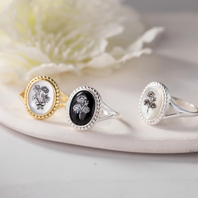 K&ouml;nnte beinhalten: Drei Ringe mit ovalen Blumendesigns. Ein Ring hat ein goldenes Band und einen wei&szlig;en Mittelteil mit einem Blumenstrau&szlig;. Der zweite Ring hat ein silbernes Band und einen schwarzen Mittelteil mit Rosen. Der dritte Ring hat ein silbernes Band und einen wei&szlig;en Mittelteil mit einem Blumendesign.