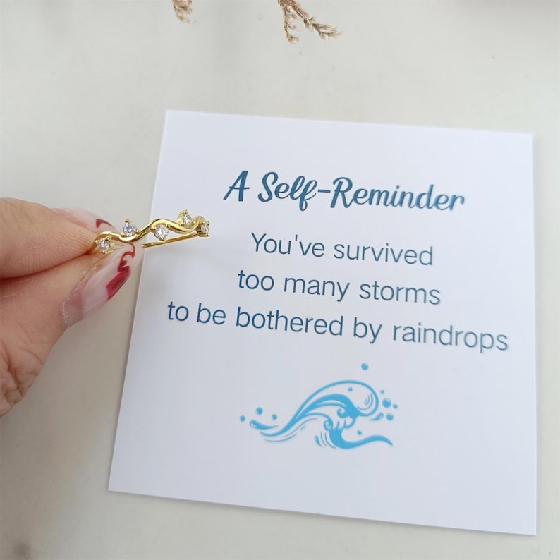 Puede incluir: Un anillo de oro con peque&ntilde;as piedras transparentes engastadas en un dise&ntilde;o de onda. El anillo est&aacute; sobre una tarjeta blanca con el texto "A Self-Reminder You've survived too many storms to be bothered by raindrops".