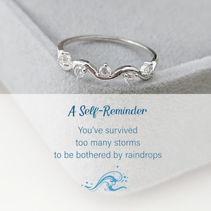 Puede incluir: Un anillo de plata con una banda ondulada y peque&ntilde;as piedras transparentes. El anillo est&aacute; en una caja blanca con el texto "A Self-Reminder You've survived too many storms to be bothered by raindrops" y un gr&aacute;fico de una ola azul.