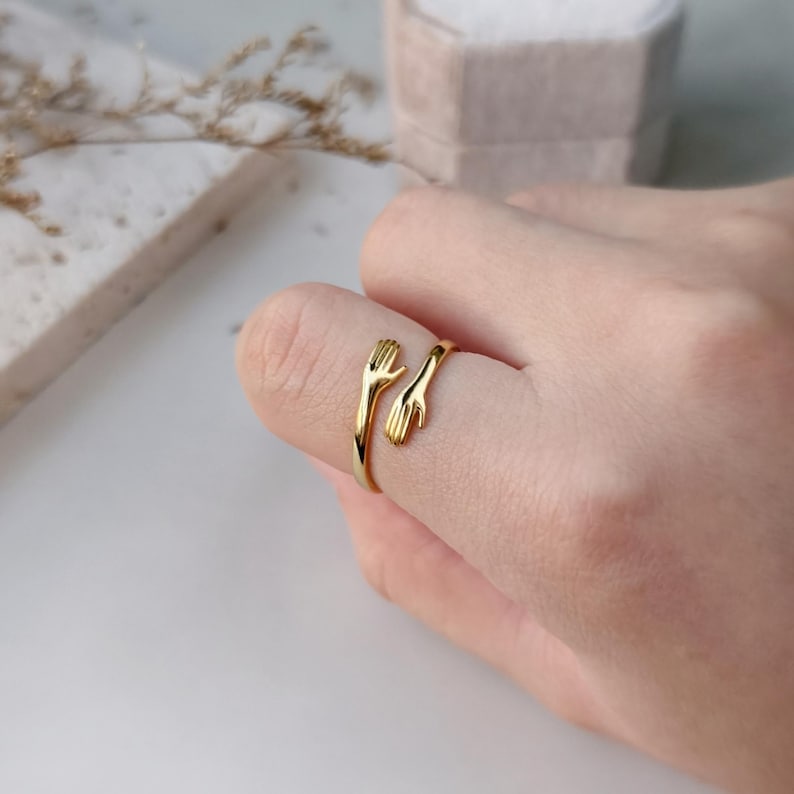 Puede incluir: Un anillo abierto de tono dorado con un dise&ntilde;o de dos manos que se extienden una hacia la otra. La banda es pulida y lisa, con las manos posicionadas para envolver el dedo, creando una apariencia continua y conectada sobre el dedo.