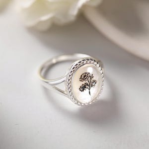 K&ouml;nnte beinhalten: Ein silberner Ring mit einem ovalen Perlmutt-Gesicht mit einem schwarzen Blumendesign. Der Ring hat einen geflochtenen Rand und ein schlichtes Band. Der Ring ist ein Schmuckst&uuml;ck.