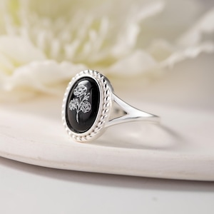 K&ouml;nnte beinhalten: Silberring mit einem ovalen, schwarzen Onyxstein. Der Stein zeigt ein wei&szlig;es Blumenmuster. Der Ring hat einen geflochtenen Rand um den Stein und ein geteiltes Schaftdesign. Der Ring liegt auf einer wei&szlig;en Oberfl&auml;che.