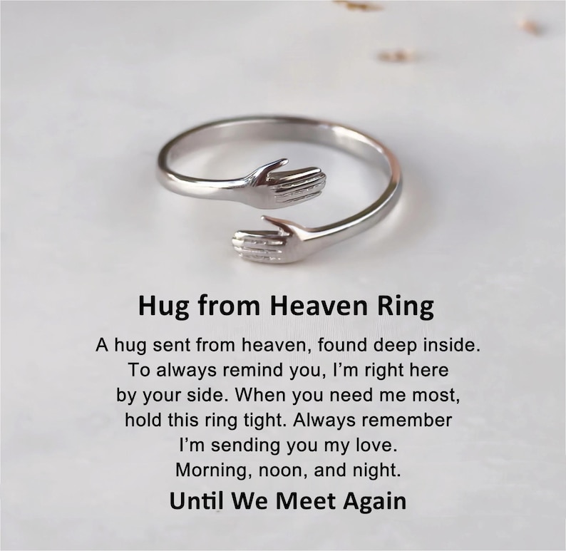 Może przedstawiać: Srebrny pierścionek z dwoma splecionymi dłońmi, tworzącymi uścisk. Pierścionek jest grawerowany słowami "Hug from Heaven Ring" i wierszem wyjaśniającym jego znaczenie.