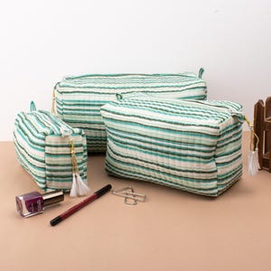 May include: Three striped green and white quilted makeup bags with gold zipper pulls and tassels. The bags are arranged in a pyramid shape on a light brown surface.