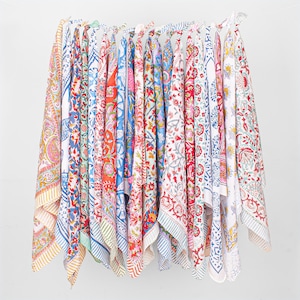 Puede incluir: Una colección de textiles coloridos estampados colgados sobre un fondo blanco. Los textiles presentan diseños florales y geométricos en tonos rojos, azules, rosas y amarillos. Las telas son probablemente de algodón o lino, adecuadas para la decoración del hogar.