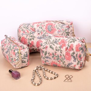 May include: Three pink and green floral print fabric zippered pouches with tassels. The pouches are different sizes and are arranged on a light brown surface.
