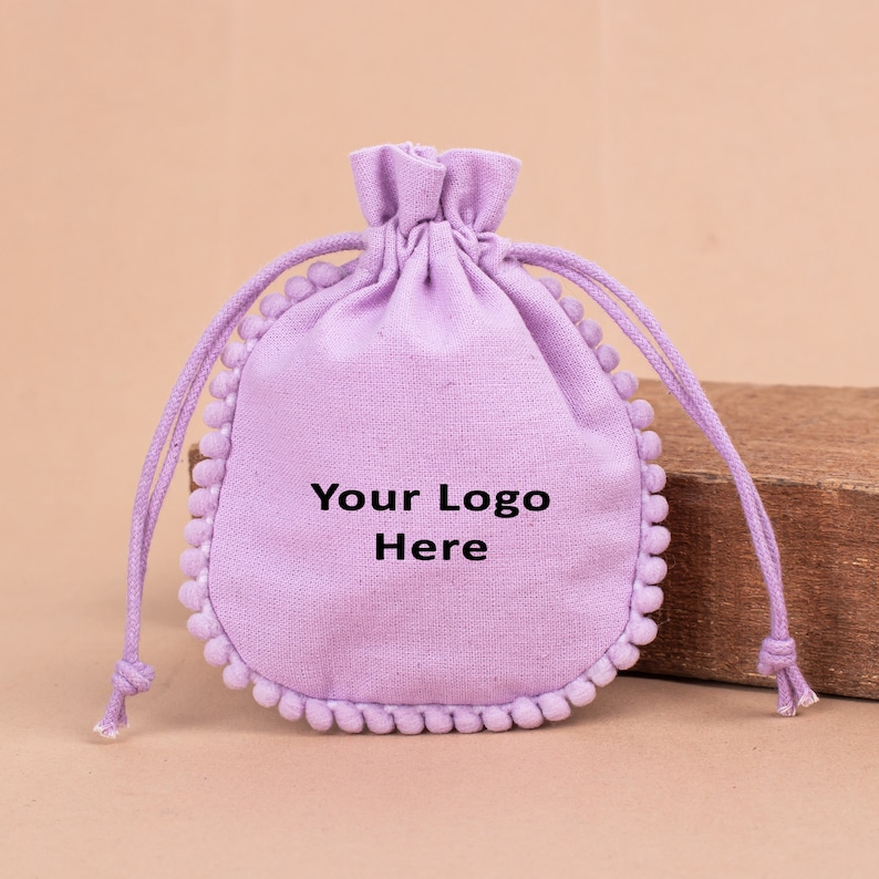 May include: A lavender drawstring pouch with a rounded bottom and a decorative pom-pom trim. The pouch has the text "Your Logo Here" printed on the front. The drawstring is made of the same lavender material.