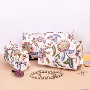 May include: Three white quilted cosmetic bags with a floral pattern in red, blue, and green. The bags are different sizes and have a zipper closure with a tassel. A bottle of nail polish and a beaded bracelet are in the foreground.