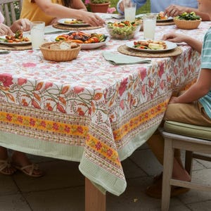 May include: A floral tablecloth with a white background and pink, orange, and green accents. The tablecloth is on a wooden table, set for a meal with plates, glasses, and food. The tablecloth has a striped border.