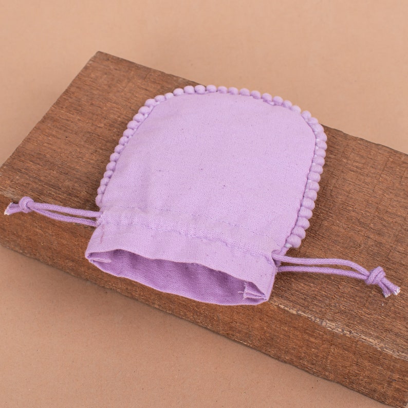 May include: A lavender drawstring pouch with a rounded top and a border of small, matching pom-poms. The pouch is made of a soft fabric and sits on a wooden surface. The drawstring is tied.