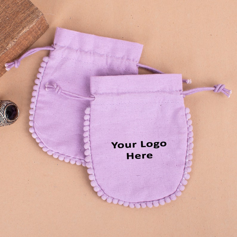 May include: Two lavender drawstring pouches with a pom-pom trim. The pouches have a rounded bottom and a drawstring closure. The text "Your Logo Here" is printed on the front of the smaller pouch. The pouches are on a beige surface.