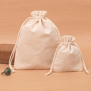 May include: Two beige drawstring bags of different sizes. The bags are made of a textured fabric and have a gathered top with a drawstring closure. The bags are sitting on a light brown surface.