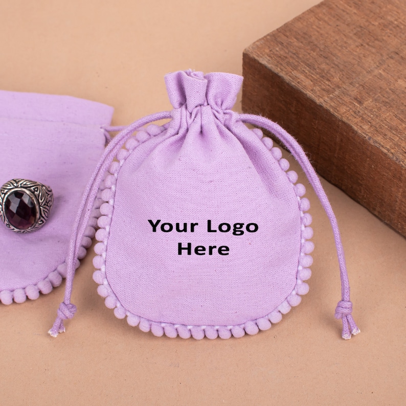 May include: Lilac drawstring pouch with white pom-pom trim and the text "Your Logo Here". A silver ring with a dark purple stone is visible. The pouch is made of fabric and has a drawstring closure.