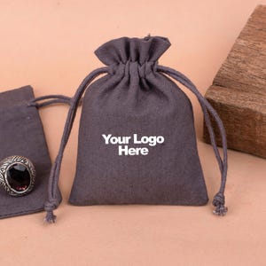 May include: A gray drawstring pouch with the text "Your Logo Here" printed on the front. The pouch is made of a soft, textured fabric and is perfect for storing small items.