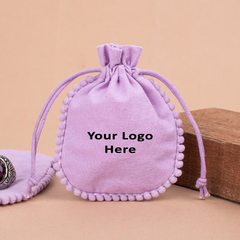 Pastel purple fabric drawstring gift pouch with pom-pom edging and custom logo space, styled with soft floral accents and neutral décor, ideal for branded packaging, jewelry, keepsakes, and small gifts.