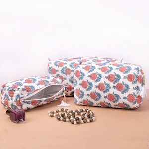 May include: A set of three white cosmetic bags with a floral pattern in coral and blue. One bag is open, revealing the interior. A bottle of nail polish and a beaded necklace are also present.