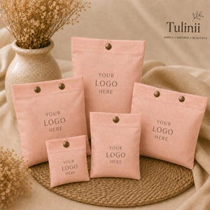 May include: A collection of five soft, dusty pink fabric pouches in varying sizes, each featuring a single metallic snap button closure and the printed text "YOUR LOGO HERE" centered on the front. The pouches are arranged in a group.