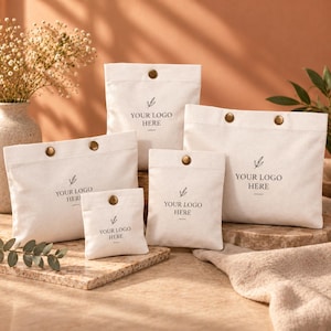 May include: A collection of four off-white canvas pouches in varying sizes, each featuring a single bronze-colored snap button closure and the printed text "YOUR LOGO HERE" accompanied by a small leaf graphic centered on the front surface.