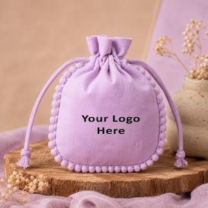 Light lavender cotton drawstring pouch with decorative pom-pom trim and customizable logo print, displayed on a rustic wooden surface with dried flowers and ceramic décor, suitable for gift packaging, jewelry storage, favors, and small accessories.