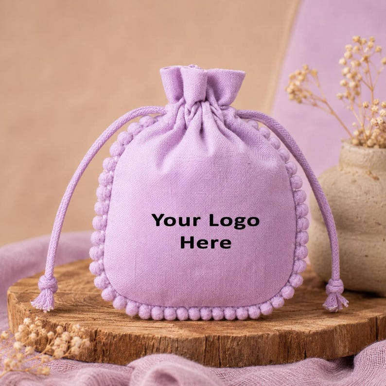 Light lavender cotton drawstring pouch with decorative pom-pom trim and customizable logo print, displayed on a rustic wooden surface with dried flowers and ceramic décor, suitable for gift packaging, jewelry storage, favors, and small accessories.