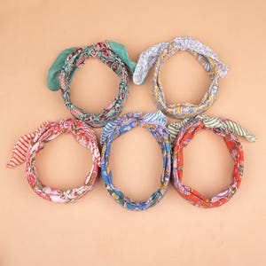 May include: Five colorful fabric headbands with bow details. The headbands feature various floral and patterned designs in shades of red, blue, green, and pink. The headbands are arranged in a circular pattern against a neutral background.