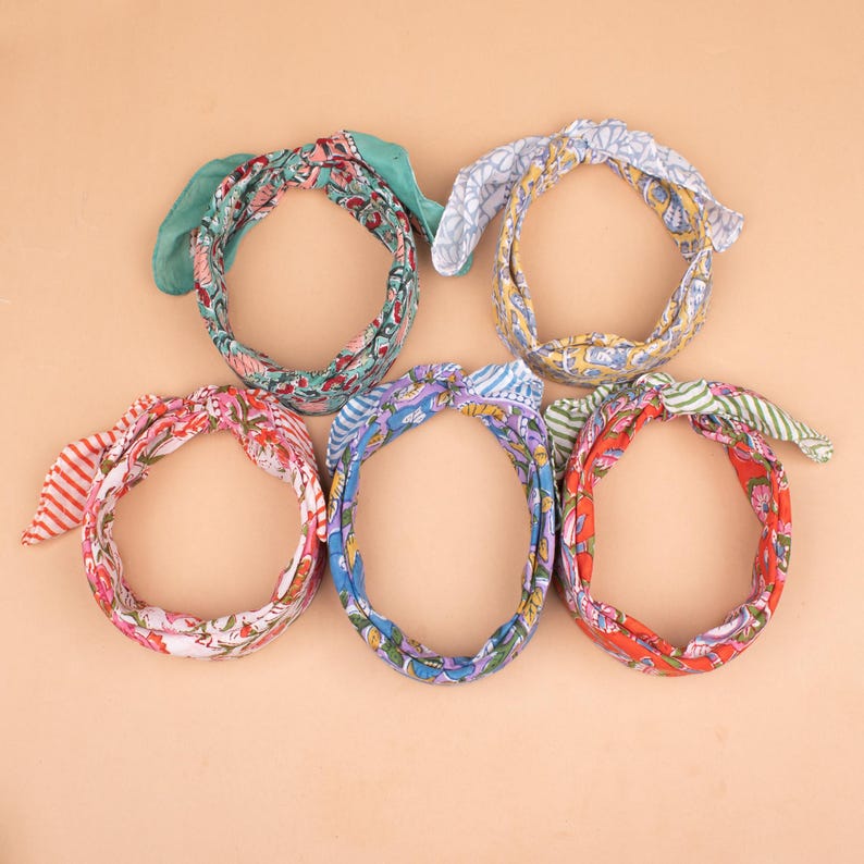 May include: Five colorful fabric headbands with bow details. The headbands feature various floral and patterned designs in shades of red, blue, green, and pink. The headbands are arranged in a circular pattern against a neutral background.