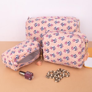 May include: Three peach-colored cosmetic bags with a floral pattern in blue and pink. The bags are different sizes and have gold zippers and tassels. A bottle of nail polish and a beaded necklace are also visible.