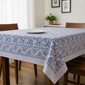 May include: A square tablecloth featuring a blue and white floral pattern with a detailed border design. The fabric displays intricate botanical motifs across the center, framed by a wide decorative band and a thin striped edge along the bottom.