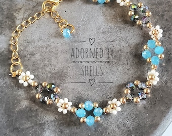 May include: A delicate gold chain bracelet with white and blue beads. The bracelet features a repeating pattern of small white flowers and blue gemstone-like beads. The clasp is a gold lobster claw.
