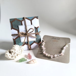 May include: A pink and white gemstone necklace with gold accents on a brown card with the text "Adorned by Shells". The necklace is packaged in a gift box with a blue and brown patterned design.