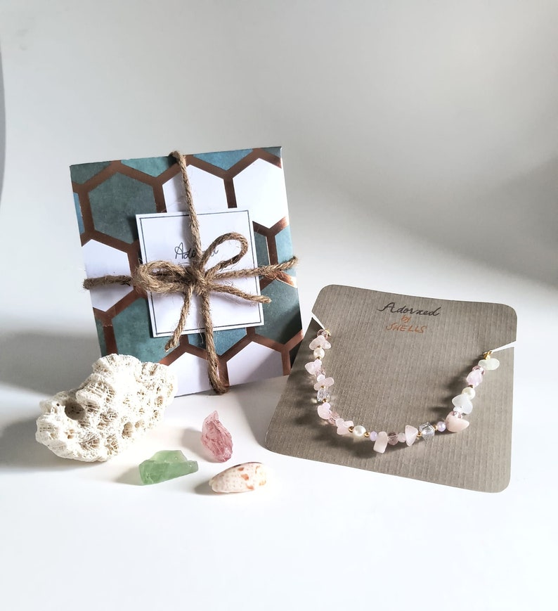 May include: A pink and white gemstone necklace with gold accents on a brown card with the text "Adorned by Shells". The necklace is packaged in a gift box with a blue and brown patterned design.