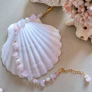 May include: A delicate bracelet made with pink and white beads, including small shell pieces, and a gold chain clasp. The bracelet is displayed on a white seashell.