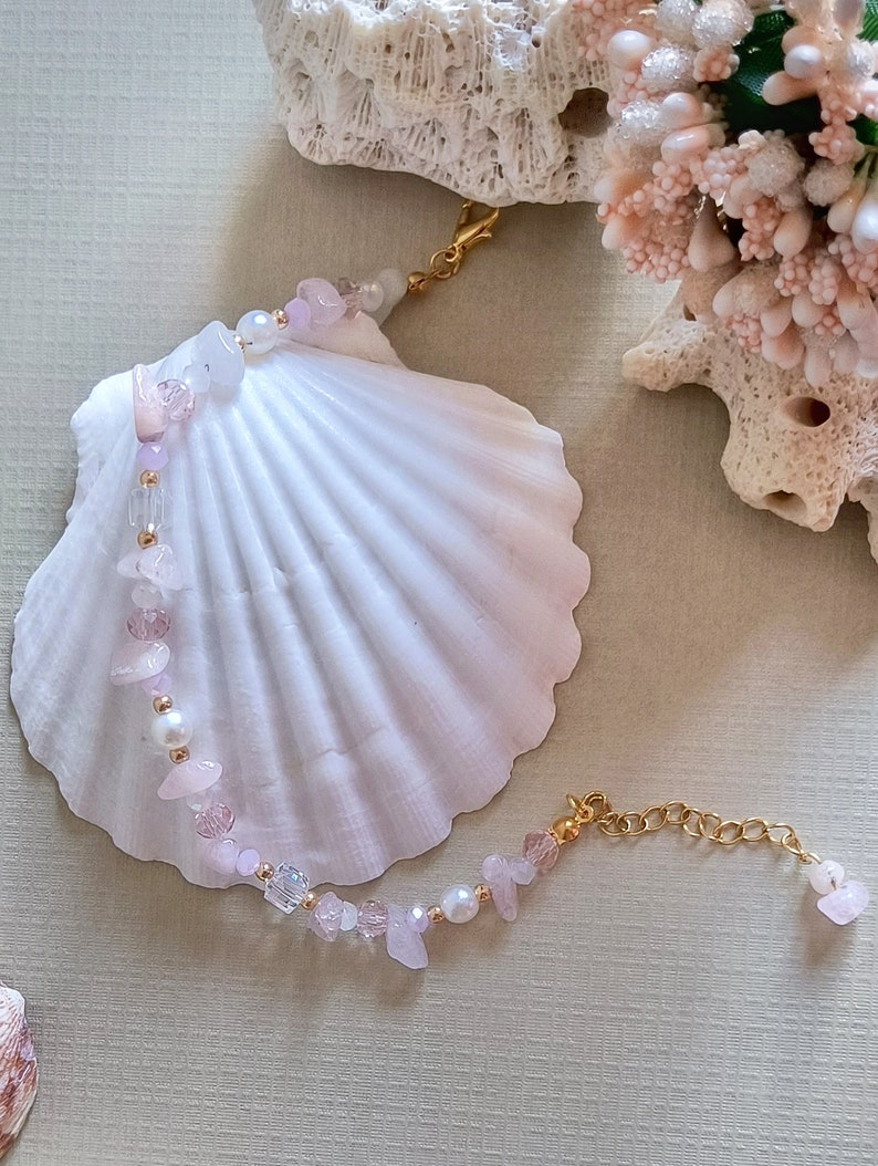 May include: A delicate bracelet made with pink and white beads, including small shell pieces, and a gold chain clasp. The bracelet is displayed on a white seashell.