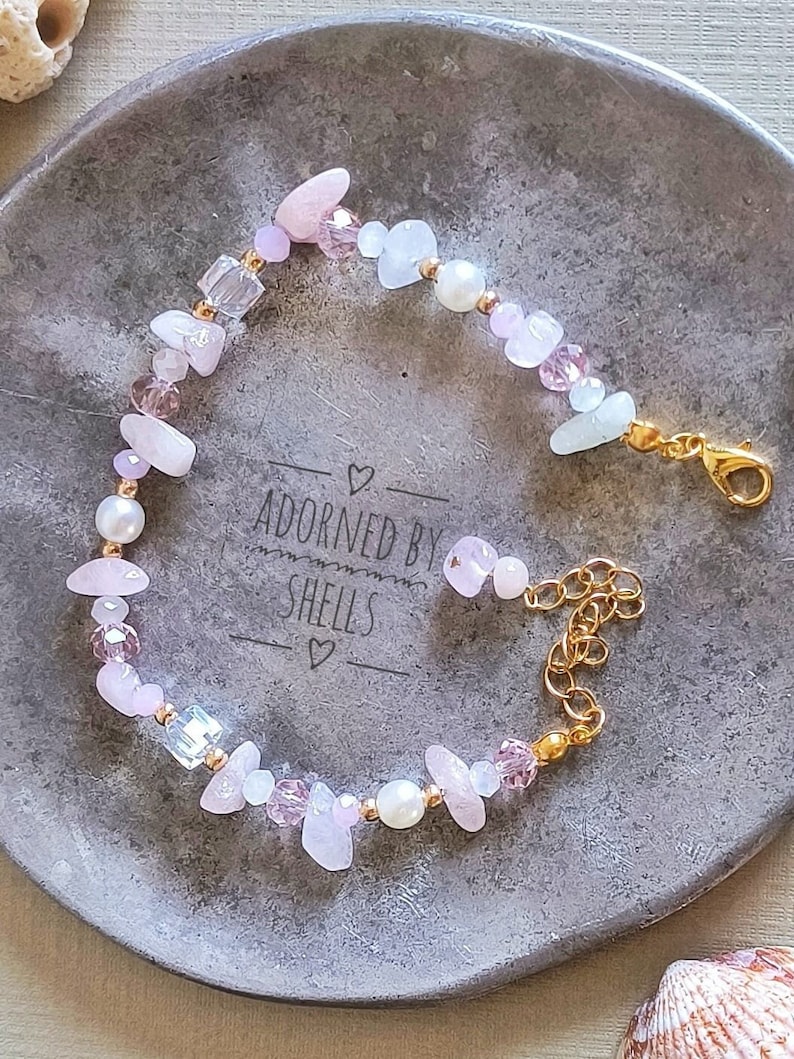May include: A delicate bracelet featuring pink and white beads, including rose quartz, pearls, and glass beads. The bracelet has a gold clasp and is adorned with the text "Adorned by Shells".