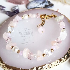 May include: A delicate pink and white gemstone bracelet with a gold clasp. The bracelet is on a white and gold decorative dish. The text "Adorned by Shells" is printed on the dish.