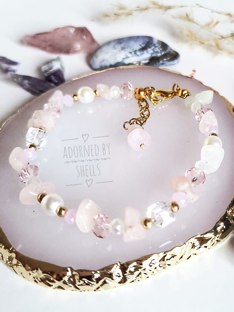 May include: A delicate pink and white gemstone bracelet with a gold clasp. The bracelet is on a white and gold decorative dish. The text "Adorned by Shells" is printed on the dish.