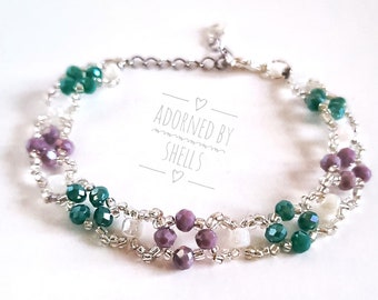 May include: A delicate silver chain bracelet with green, purple, and white beads arranged in a floral pattern. The bracelet features a clasp closure and the text "Adorned by Shells" is printed on a white background.